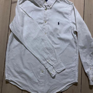 Vit skjorta från Ralph Lauren - Klassisk vit skjorta från Ralph Lauren med knappar framtill och button-down krage. Skjortan är långärmad och har en stilren design som passar till många tillfällen. Tillverkad i bomull för en bekväm känsla. Perfekt till skolavslutningen 