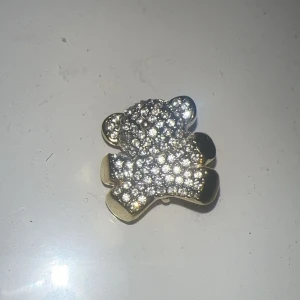 Guldfärgad björn brosch med strass - Söt brosch i form av en björn, täckt med glittrande strass på framsidan och guldfärgad baksida. Perfekt accessoar för att ge din outfit lite extra bling och personlighet.