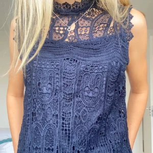 Marinblå spetsklänning med hög hals - Säljer en marinblå klänning med vacker spets och hög hals. Klänningen är ärmlös och har fina detaljer i spetsen upptill. Perfekt för dig som vill ha en elegant och stilren look.