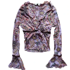 Vintage Mönstrad topp i mesh med volanger - Säljer en lila och rosa omlottopp i mesh med paisleymönster och volangdetaljer. Toppen har långa ärmar med utsvängda ärmslut och knyt framtill. Perfekt för dig som gillar färg och mönster!