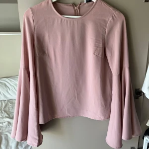Ljusrosa blus med vida ärmar från NA-KD - Säljer en ljusrosa blus från NA-KD x Hannalicious i storlek 34. Blusen har rund halsringning och långa, vida ärmar som ger en snygg och trendig look. Perfekt att styla med jeans eller kjol.