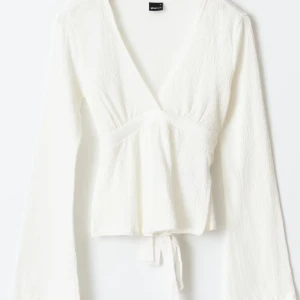 Vit omlottopp med knytband - Säljer en vit omlottopp med v-ringning och långa ärmar. Toppen har knytband i midjan och är tillverkad i ett lätt, strukturerat material. Perfekt för en stilren och enkel look. Från Gina tricot, säljer då den är lite förstor för mig. 