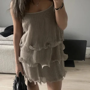 Volang klänning  - Säljer en beige plisserad klänning från H&M med volangdetaljer i flera lager. Klänningen är ärmlös med smala axelband och har en luftig, lös passform. Perfekt för varma dagar och somriga tillfällen. 
