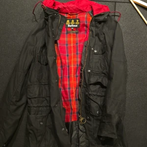 Svart jacka från Barbour med röd-rutigt foder - Klassisk svart Barbour-jacka med röd-rutigt innerfoder och guldfärgad dragkedja. Jackan har flera fickor framtill, huva med röd insida och detaljer som tryckknappar och Barbour-logga på dragkedjan.