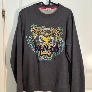 Grå sweatshirt med tiger från Kenzo - Grå sweatshirt från Kenzo med ikoniskt broderat tigerhuvud i flera färger på bröstet. Rund halsringning och ribbade muddar. Perfekt statement-plagg för dig som gillar färg och mönster.