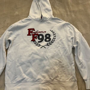 Vit hoodie från Firefly - Vit hoodie från Firefly med tryck på ryggen: 'FF 98' i rött och svart samt en lagerkrans. Klassisk huva och lång ärm, perfekt för en avslappnad stil.
