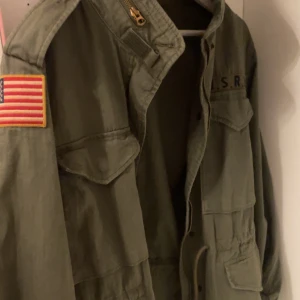  Polo Ralph Lauren safarijacka  - Säljer en grön militärinspirerad jacka från Polo Ralph Lauren med amerikansk flagga på ärmen och flera fickor framtill. Jackan har klassisk krage, dragkedja och knappar samt broderad detalj på bröstet.