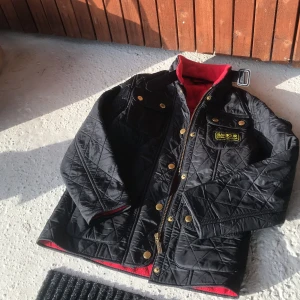 Svart quiltad jacka från Barbour - Säljer en svart quiltad jacka från Barbour International med gulddetaljer och dragkedja. Jackan har lång ärm, flera fickor framtill och röd fleecefodring på insidan. Klassisk design med Barbour-logga på bröstet barn Jacka tveka inte att höra av dig vid frågor 😉