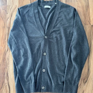 Mörkgrå kofta från jack and jones - Säljer en stilren mörkgrå kofta från jack and jones med v-ringning och knäppning framtill. Tröjan har långa ärmar och ribbade muddar vid ärmslut och nederkant. Perfekt för lager-på-lager och enkel att matcha.