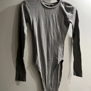 Grå långärmad bodysuit från Zara - Säljer en enkel och stilren grå långärmad bodysuit från Zara. Perfekt att styla med jeans eller kjol för en clean look. Mjuk bomullskänsla och rund halsringning.