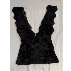 Svart blommig spets topp med djup v-ringning - Säljer en svart topp med blommigt sammetsmönster och djup v-ringning både fram och bak. Toppen har spetsdetaljer längs kanterna och är ärmlös. Perfekt för att skapa en snygg och trendig look.