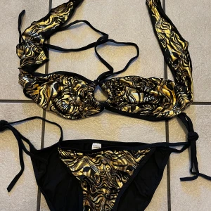 Svart och guld bikini med knyt - Säljer en snygg bikini från H&M i svart och guld med abstrakt mönster. Både överdel och underdel har knytband i sidorna och nacken. Materialet är syntet och bikinin har en glansig finish som ger en lyxig känsla.