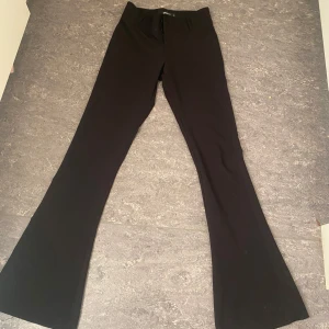 Svarta bootcut kostymbyxor med fickor - Snygga svarta kostymbyxor med bootcut-ben och två bakfickor med knappar. Byxorna har klassisk passform och stängs med dragkedja och knapp framtill. Perfekta för en stilren look. 
