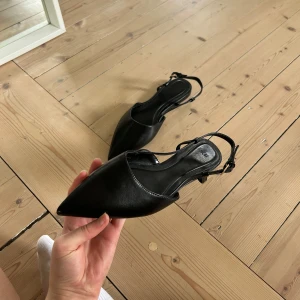 Svarta slingback pumps med spetsig tå - Säljer ett par svarta pumps från H&M med smala slingback-remmar och spetsig tå. Skorna har en elegant design med liten klack och justerbart spänne vid ankeln. Perfekta för dig som gillar stilrena och klassiska skor.