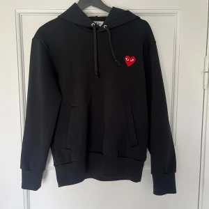Svart hoodie från Comme des Garçons Play - Svart hoodie från Comme des Garçons Play med den ikoniska röda hjärtloggan på bröstet. Tröjan har huva med dragsko och fickor framtill. Perfekt för en stilren och trendig look.