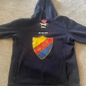 Blå hoodie från CCM med DIF-logga - Mörkblå hoodie från CCM med stor DIF-logga på bröstet och snörning vid halsen. Tröjan har huva och långa ärmar, perfekt för dig som vill visa ditt stöd för Djurgården. Klassisk sportig stil.