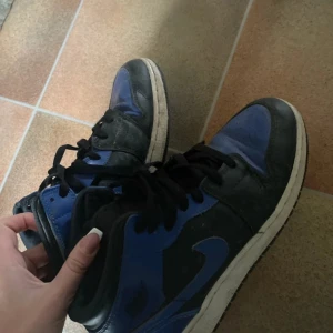 Nike Air Jordan 1 Retro High OG blå och svarta sneakers - Säljer ett par Nike Air Jordan 1 Retro High OG sneakers i blått och svart med klassisk snörning och rund tå. Skorna har den ikoniska Air Jordan-loggan på sidan och är perfekta för dig som gillar streetwear och sneakers.