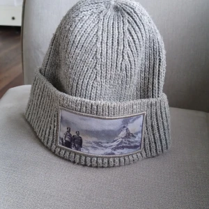 Grå ribbstickad mössa med patch - Grå ribbstickad mössa med bred uppvikt kant och en rektangulär patch framtill som visar ett motiv av två personer och ett berg. Mössan är mjuk och passar perfekt för kyligare dagar. Mössan är från revenite AB och är limited edition för den säljs inte längre. Pris kan diskuteras 