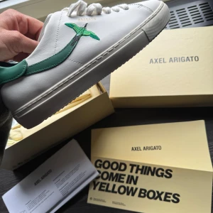 Vita sneakers från Axel Arigato med grön fågel - Snygga vita sneakers från Axel Arigato med en grön fågeldetalj. Sprillans nya endast provade, säljer endast för dom inte satt bra på min fot. Skickas i Orginal kartong. Ute ur sortiment på hemsidan nypris 2750 