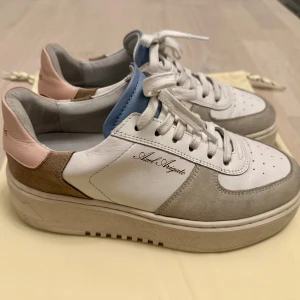 Axel Arigato orbit sneakers i vitt, grått och rosa str 38 - Snygga sneakers från Axel Arigato i vitt skinn med grå mockadetaljer och ljusrosa häl. Skorna har vita snören, blå insida vid plösen och en tjock platt sula. Märkeslogga på sidan och bak på hälen.Perfekt för en avslappnad men trendig look.