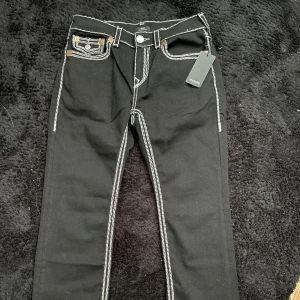 Svarta jeansbyxor från True Religion med vit kontrastsöm - Svarta jeansbyxor från True Religion med tydliga vita kontrastsömmar och dekorativa fickor bak. Klassisk femficksmodell med knappstängning och logotyp på bakfickan. Snygg detalj med märkets emblem på bakfickan.