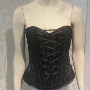 Svart korsett med snörning (ej underklädeskorsett) M - Snygg svart korsett med dekorativa snörningar både fram och bak. Axelbandslös modell med markerad midja och lätt glansigt tyg. Perfekt för att skapa en edgy look. Helt ny. Storlek M.