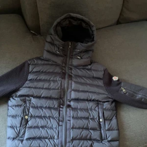 Svart pufferjacka från Moncler - Säljer en svart pufferjacka från Moncler med stickade ärmar och dragkedja framtill. Jackan har hög krage, huva med snörning och två fickor med tryckknappar. Moncler-logga på ena ärmen och detaljer i både syntet och stickat material.