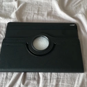 Svart fodral till IPad Pro 9,7 med roterande stativ - Säljer ett svart fodral till surfplatta med roterande stativfunktion. Fodralet har en rund öppning på baksidan och är gjort i ett material som liknar skinn. Perfekt för att skydda din surfplatta och använda den i olika vinklar.