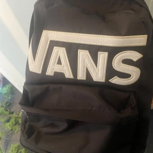 Svart ryggsäck från Vans - Säljer en svart ryggsäck från Vans med stort vitt broderat logotyp på framsidan. Väskan har ett stort huvudfack och en mindre ficka framtill med dragkedja. Perfekt för skolan eller vardagsbruk.
