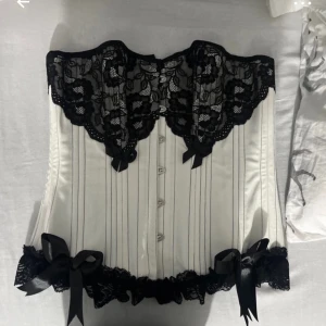 Vit korsettopp med svarta spetsdetaljer och rosetter - Säljer en elegant vit korsettopp med svarta spetsdetaljer upptill och nedtill samt dekorativa svarta rosetter. Toppen har snörning i ryggen och är axelbandslös, perfekt för att skapa en snygg siluett. (Mamma sydde den fast tyvörr va den för stor för mig🥲) diskutera priset vid snabb affär