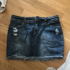 Jeanskjol med slitningar från ONLY - Mörkblå jeanskjol från ONLY med slitna detaljer framtill och bakfickor. Kjolen har rå kant nertill och klassisk femficksdesign. Aldrig använd