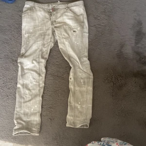 Grå jeansbyxor från Dsquared2 - Säljer ett par grå jeansbyxor från Dsquared2 i storlek 50. Byxorna har en tvättad look med vita färgstänk och slitningar, klassisk femficksmodell och knappar i gylfen. Perfekta för dig som gillar streetwear och vill sticka ut. PRIS DISKUTERAS