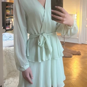 Ljusgrön omlottklänning med volang och knytband - Säljer en ljusgrön omlottklänning med lång ärm och knytband i midjan. Klänningen har volangdetaljer vid midjan och en mjuk, flowy känsla. Perfekt för vår och sommar, med en feminin och fräsch look.