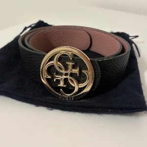 Svart bälte med guldspänne från Guess - Snyggt svart bälte från Guess med ett stort, rundat guldspänne med logotyp. Tygpåse medföljer. Meddela för fler bilder. 