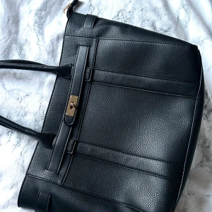 Svart handväska med gulddetalj - Snygg svart handväska i skinnimitation med stilren design och guldfärgad låsdetalj framtill. Väskan har dubbla handtag och rymligt huvudfack, perfekt för att bära med sig det viktigaste till vardags.