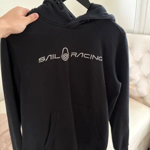 Svart hoodie från Sail Racing - Säljer en svart hoodie från Sail Racing med vit logga broderad på bröstet. Tröjan har huva och en stor magficka framtill. Perfekt för dig som gillar en sportig och stilren look.