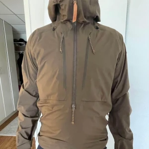 Brun vindjacka från Fjällräven med huva - Säljer en brun vindjacka från Fjällräven med justerbar huva och dragkedjor framtill. Jackan har snygga detaljer som snörning vid huvan och klassisk Fjällräven-logga på ärmen. Perfekt för utomhusaktiviteter och stilren look.