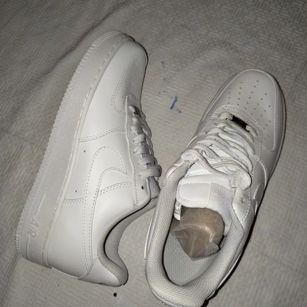Säljer ett par klassiska helvita Nike Air Force 1 sneakers. Skorna har en stilren design med perforerad tå, snörning och platt sula. Perfekta för dig som gillar en clean och tidlös look. Materialet är skinn och de passar till många olika outfits.. Kengät.