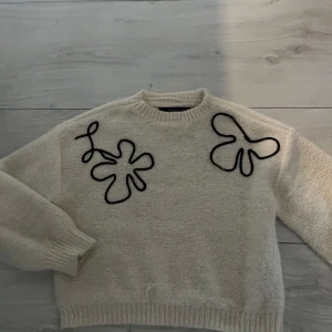 Beige stickad tröja med svart broderi - Säljer en beige stickad tröja med unika svarta broderade blommor på bröstet. Tröjan har rund halsringning och långa ärmar med ribbade muddar. Perfekt för dig som vill ha något annorlunda och mysigt i garderoben!