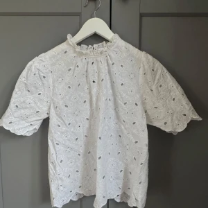 Vit blus med broderade detaljer från Holly & Whyte - Superfin vit blus från Holly & Whyte med broderade hålmönster och volang vid halsen. Blusen har korta puffärmar och en söt knapp i nacken. 