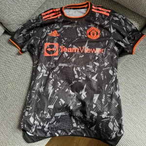 Manchester United bortatröja 2022-23 - Manchester Uniteds bortatröja från säsongen 2022/23. Svart bas med orange detaljer, adidas-logga och TeamViewer-tryck. Unik grafisk design över hela tröjan ger den en modern och sportig look. Väldigt skön att ha på sig tack vare det lätta materialet. Perfekt både till match, träning eller som en snygg streetwear-tröja
