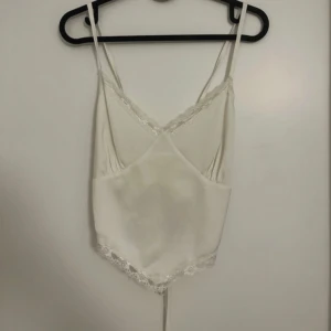 Vit spetsdetaljerad linnetopp med snörning - Säljer en vit linnetopp med tunna axelband och spetsdetaljer längs urringningen och nederkanten. Toppen har v-ringning och snörning i ryggen för en justerbar passform. Perfekt för sommardagar eller som ett lagerplagg.