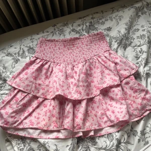 Rosa blommig volangkjol från Gina Tricot - Supersöt rosa kjol med små blommor från Gina Tricot. Kjolen har två lager volanger och en bred, smockad midja som ger en bekväm passform. Perfekt för dig som gillar romantisk och lekfull stil. Passar mig som är 160!