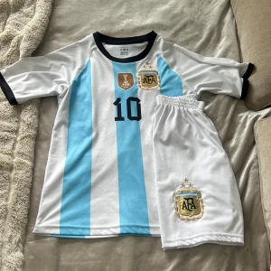 Argentina Messi 10 fotbollströja och shorts - Säljer ett set med Argentina fotbollströja och matchande shorts. Tröjan har klassiska blå och vita ränder, svart krage och ärmar, nummer 10 och Messi-tryck på ryggen. Både tröja och shorts har AFA-emblem och guldiga detaljer.