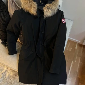 Svart parkajacka med päls från Canada Goose - Säljer en svart parkajacka från Canada Goose med stor huva kantad av ljus päls. Jackan har knappar framtill och klassisk logga på ärmen. Perfekt för kalla dagar.