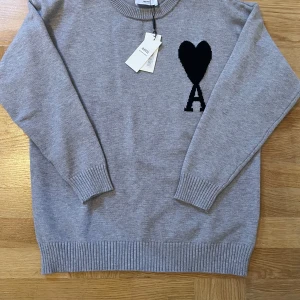 Grå stickad tröja från Ami Paris - Säljer en grå stickad tröja från Ami Paris med svart hjärta och bokstaven A broderat på bröstet. Tröjan har rund halsringning och långa ärmar med ribbade muddar. Perfekt för en stilren och trendig look.