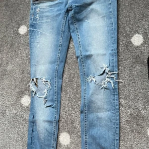 Blå slitna jeans  Gina tricot - Säljer ett par blå jeans från Kristen med slitningar och hål på båda benen. Jeansen har klassisk femficksdesign och normal passform. Perfekta för en avslappnad och trendig look.