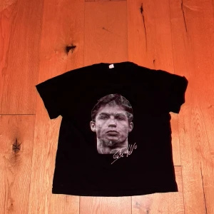 Svart t-shirt med Ronaldo-tryck - Svart t-shirt med stort porträtt av fotbollsspelaren Ronaldo på framsidan och en bild av honom med nummer 7 och texten 'THE GOAT' på baksidan. Perfekt för fotbollsfans som vill visa sitt stöd.