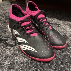Svarta och rosa fotbollsskor från Adidas - Säljer ett par svarta och rosa fotbollsskor från Adidas med vita ränder på sidan. Skorna har snörning och mönstrad sula för bra grepp på konstgräs eller grus. Perfekta för dig som vill sticka ut på planen!