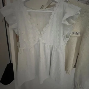 Ljus ljus blå med spets kant! - Söt vit blus med v-ringning, spetsdetaljer och volangärmar. Blusen har ett broderat mönster och är tillverkad i ett lätt bomullstyg. Perfekt för vår och sommar!